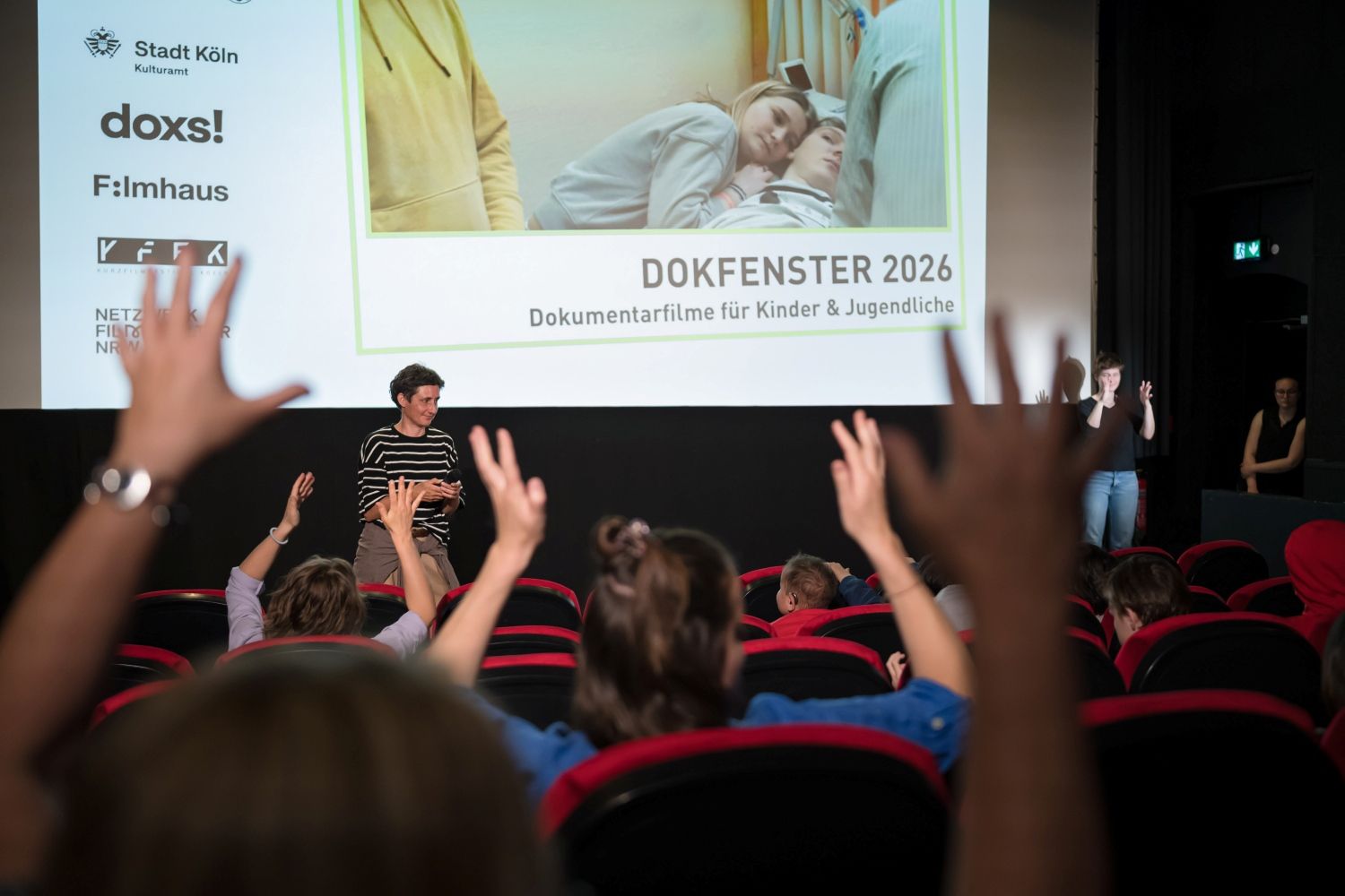 Ein Kinosaal mit roten Plüschsesseln von hinten mit Blick auf die Leinwand. Viele Kinder winken mit erhobenen Händen (Geste für Applaus). Vor der Leinwand steht eine Frau in geringeltem Oberteil mit mittelllangen dunklen Haaren. rechts von der Leinwand stehen zwei Frauen, eine gebärdet. Auf der Leinwand sehen wir ein Still des Films "Seht ihr mich?" von Leah Meinhof. Farauf kuschelt sich ein etwa 14jähriges Mädchen an einen Jungen der im Krankenhausbett liegt. darunter steht Dokfenster 2026. Dokumentarfilme für Kinder und Jugendliche. links neben dem Bild sind die Logos von Stadt Köln - Kulturamt, doxs! Filmhaus und KFFK zu sehen.