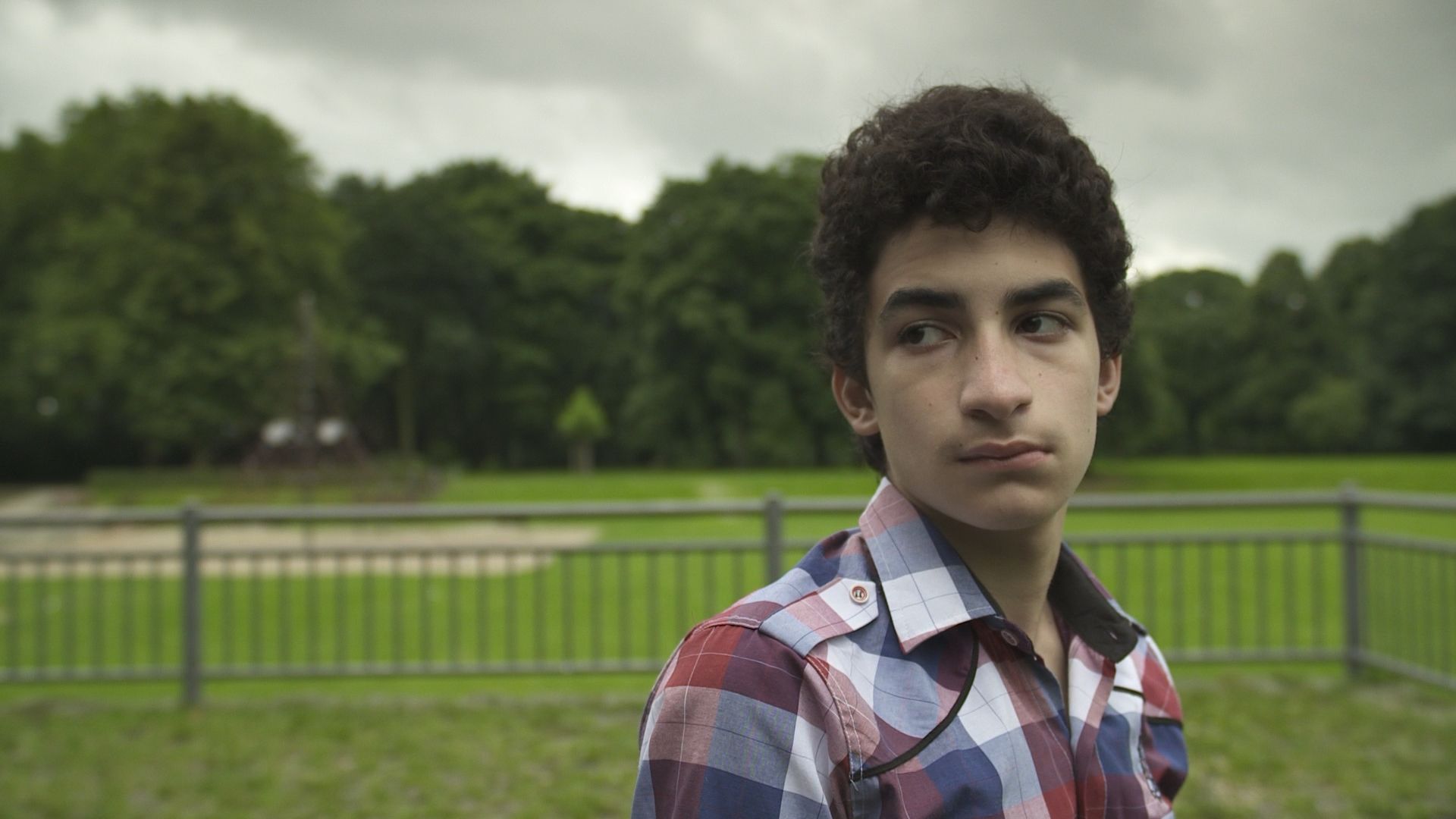 Ein junger Mann mit lockigem Haar und kariertem Hemd steht draußen in einem Park vor einem Metallzaun. Sein Blick ist ernst und nachdenklich.